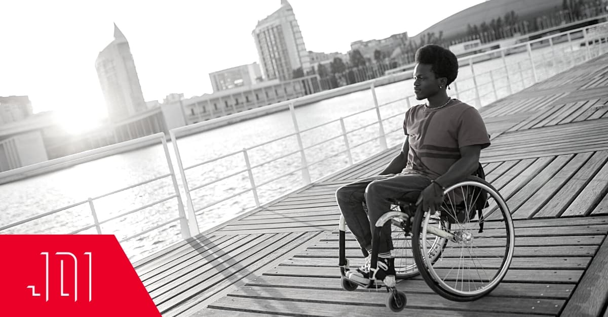 A Black masculine person in a wheelchair on a boardwalk looks out over a river. There's a cityscape behind him